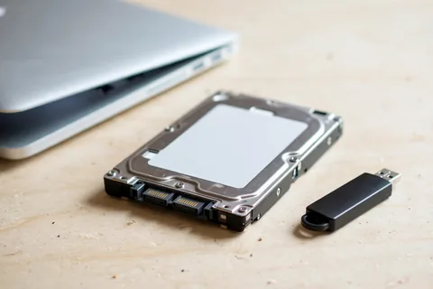 A bare 2.5-inch laptop hard drive and a black USB flash drive lying unplugged on a wooden desk beside a closed silver laptop