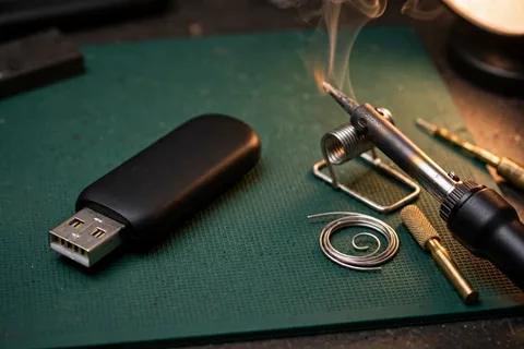 A black USB flash drive lying on a green workbench mat next to a soldering iron resting in its stand, a coil of solder wire and a brass tip cleaner