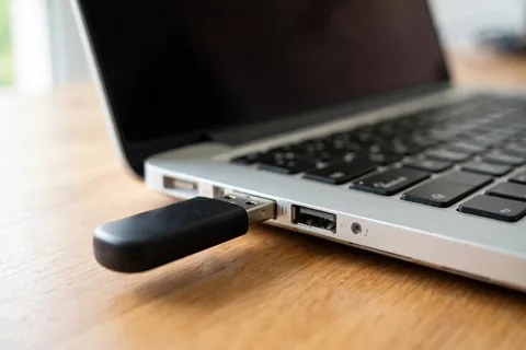 A black USB flash drive plugged into the left USB port of a silver laptop on a wooden desk