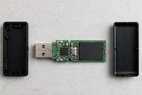 A USB flash drive taken apart on a grey desk, with the two empty case halves and the green circuit board that carries the plug, a small square controller chip, a silver crystal oscillator and a larger flat NAND memory chip