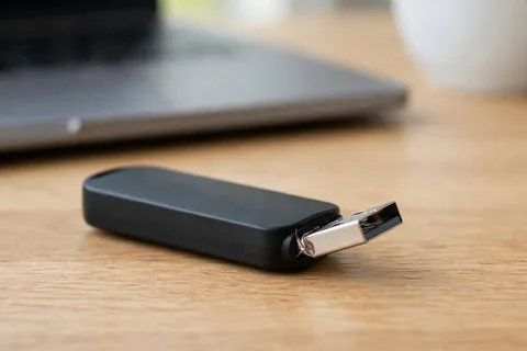 A black USB flash drive lying on an oak table with its silver metal plug bent and creased where it meets the plastic body